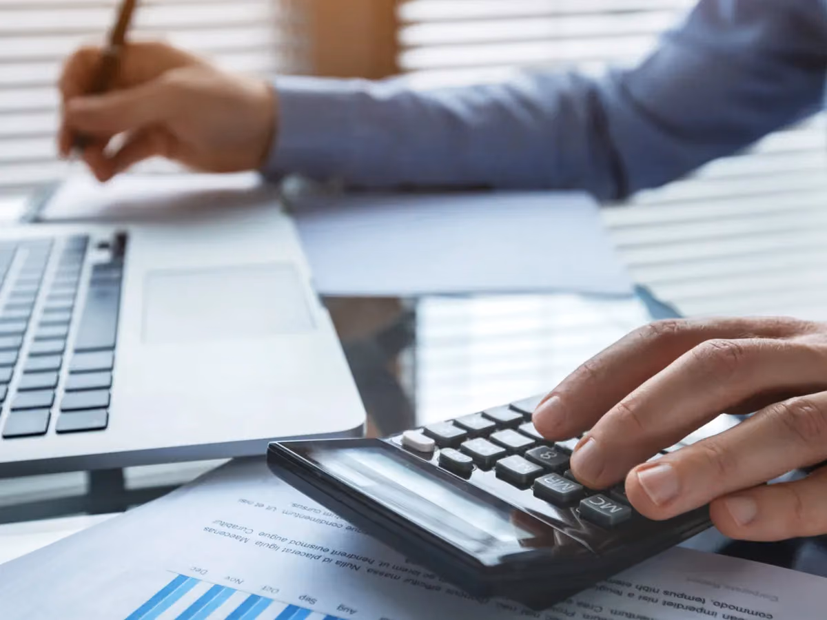 Hände an Taschenrechner und Laptop über einem Finanzbericht
